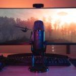 Microphone professionnel pour streaming de jeux vidéo en direct, configuré pour une qualité audio optimale, avec un moniteur de gaming et un clavier rétroéclairé, idéal pour les amateurs de jeux vidéo sur PC.