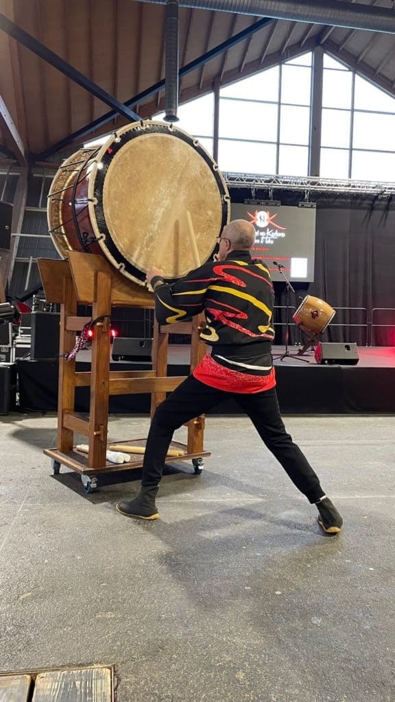 Clou de percussion géant en bois, utilisé dans un événement de musique live ou spectacle, avec un musicien qui le frappe, dans une salle avec scène et écrans lumineux.