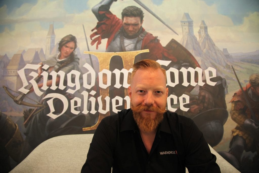 Fichier : homme-assis-devant-affiche-jeu-Kingdom-come-Deliverance.jpg.