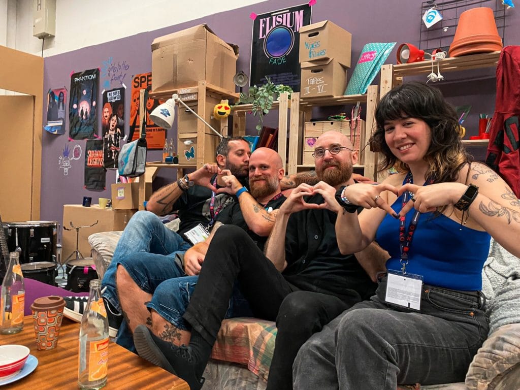Une groupe de personnes à une convention de jeux vidéo, faisant des cœurs avec leurs mains, entourés de décorations colorées et affiches de jeux sur un mur violet, représentant un espace de convivialité gaming.
