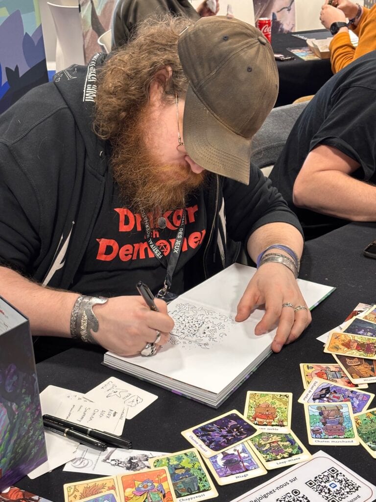 Créateur de jeux en train d'illustrer une carte dans un style de tarot fantastique lors d'une convention dédié au gaming et aux jeux de société, avec des cartes colorées et un univers immersif.