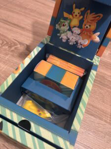 Cartes à collectionner Pokémon dans une boîte de jeu Air-Gaming, avec personnages emblématiques comme Pikachu, Flambino, et Petit Critter, pour collectionneurs et fans de Pokémon.