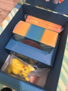 Cartes Pokémon dans une boîte de collection, comprenant un deck de cartes à jouer Pokémon et une figurine Pikachu en plastique, pour les fans de jeux de cartes et d'univers Pokémon.