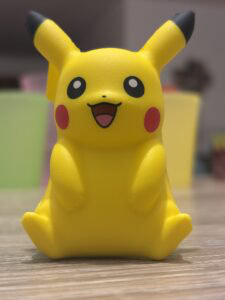 Pikachu en figurine jaune, personnage emblématique de Pokémon, exposée sur une surface en bois avec un fond flou coloré. Parfait pour jeux vidéo, collection, objets de fans, et thèmes de culture pop.