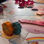 Chevalets en bois et pièce du jeu de société "Air-Gaming" avec un thème féerique, comprenant des nuages, arcs-en-ciel et éléments colorés pour une expérience ludique et imaginative.