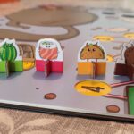 Figurines de personnages mignons en bois coloré sur une extension de jeu de société, illustrant un univers fantastique et ludique pour enfants et familles.