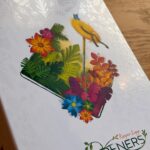 Fichier noir et blanc d'un jardin de fleurs colorées avec des plantes vertes, un chapeau de paille et une pelle sur un plateau, illustrant le jeu de société "Gardeners" d'AM. Ooms & Anthony Moulins.
