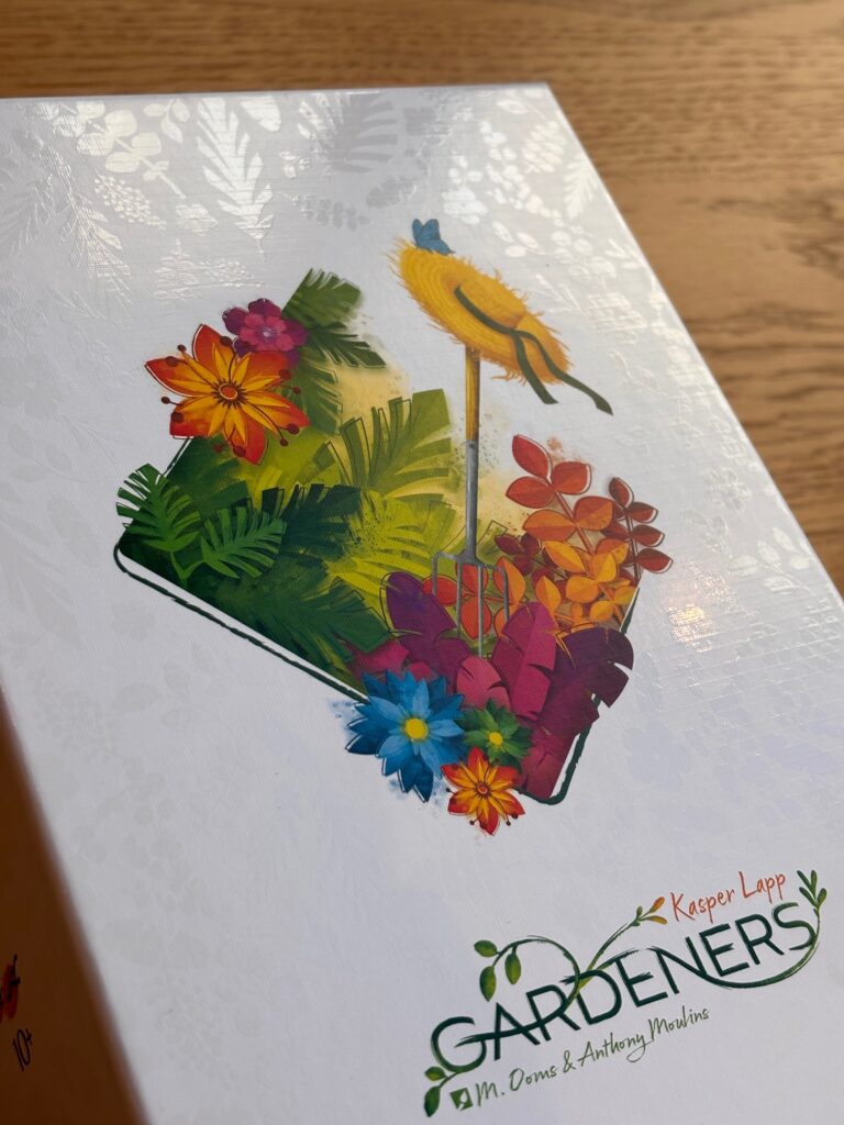 Fichier noir et blanc d’un jardin de fleurs colorées avec des plantes vertes, un chapeau de paille et une pelle sur un plateau, illustrant le jeu de société "Gardeners" d'AM. Ooms & Anthony Moulins.