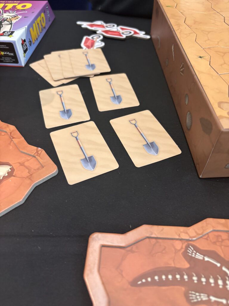 Pelle de jeu de société avec des images de pelles sur des cartes en bois, destiné à un jeu de stratégie ou de placement, avec un décor thématique de désert ou d'aventure.