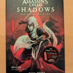 Une couverture illustrée d'une femme ninja en posture d'attaque, avec un fond sombre et rouge, représentant l'univers de jeu vidéo Assassin's Creed Shadows, édition française.