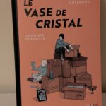 Vase de cristal, illustration de Astrid Goldsmith, avec des boîtes empilées et des objets personnels, évoquant la mémoire et la famille.