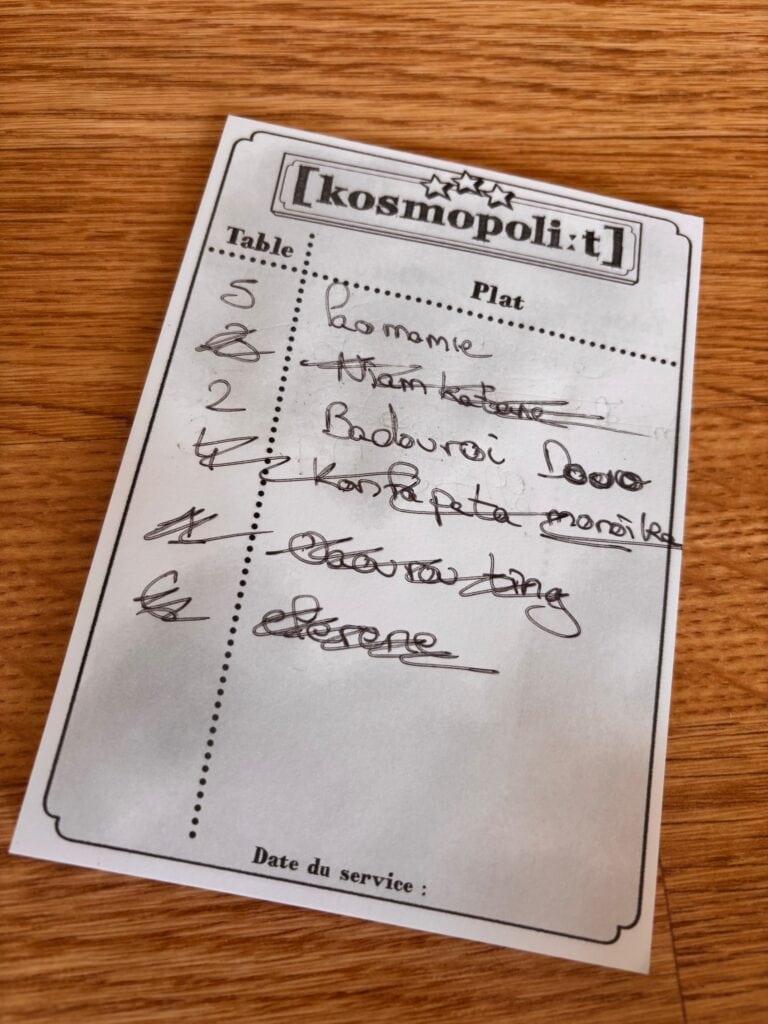 Jeu de société coopératif Kosmopoli:t, où les joueurs gèrent un restaurant sous pression, avec une liste de plats à servir.