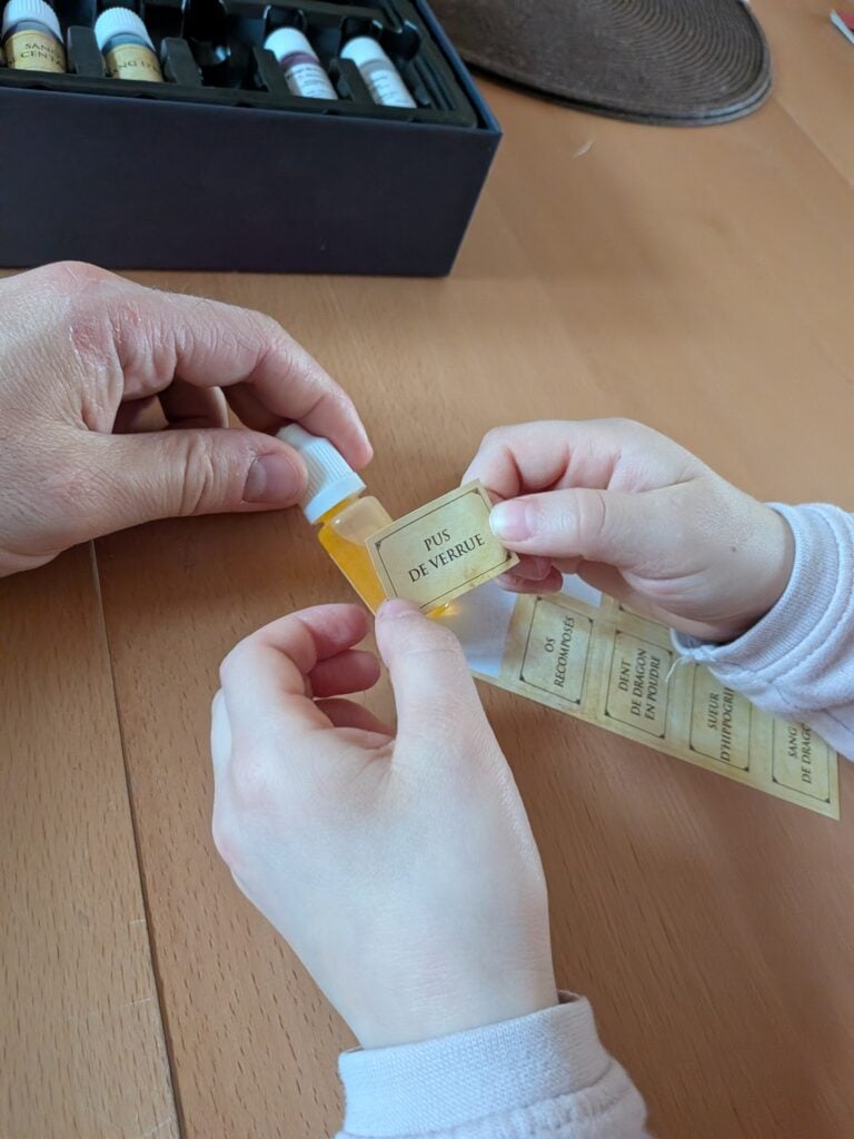 Deux mains, adulte et enfant, manipulant une petite bouteille de remède naturel, avec une boîte de flacons en arrière-plan, sur une table en bois.