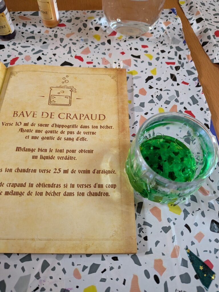 Potion verte de couleur vive dans un verre, posée à côté d’un livre ancien avec une recette de potion magique, sur une table à motifs colorés.