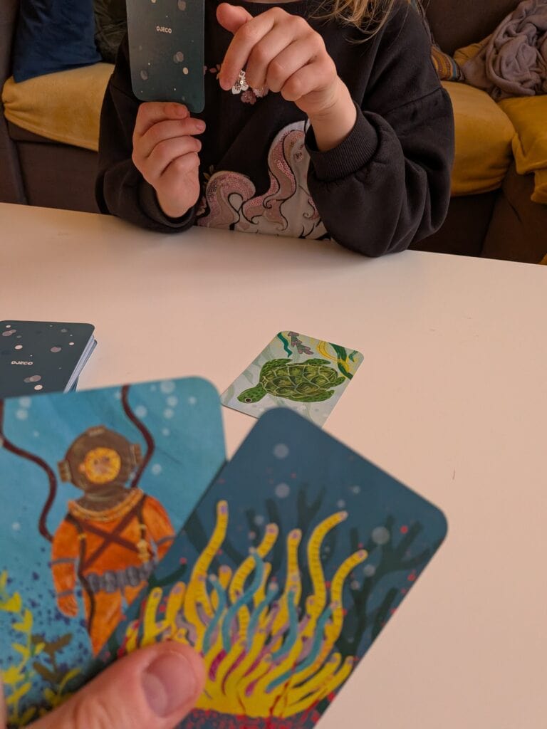 Une personne joue à un jeu de cartes avec des illustrations marines colorées, mettant en valeur un thème sous-marin avec des créatures et des éléments aquatiques.