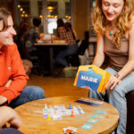 Deux femmes jouent à un jeu de société dans un café, avec un décor chaleureux et convivial, illustrant un moment de détente et de socialisation.
