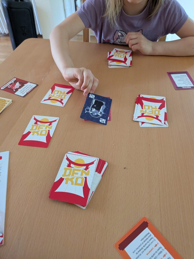 Enfant jouant à Dénko, jeu de cartes stratégique pour enfants, sur une table en bois, avec plusieurs cartes colorées et un environnement familial.