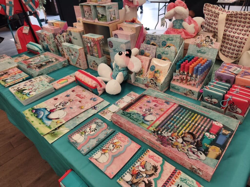 Collection de fournitures scolaires, jouets et accessoires pour enfants, comprenant des cahiers, stylos, peluches et boîtes à lunch, exposés sur une table lors d'un marché ou salon.