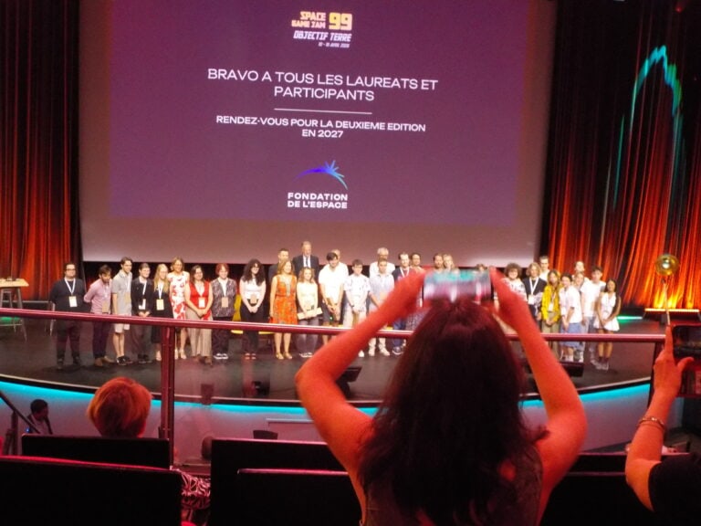 Groupe de lauréats et participants lors de la cérémonie de remise des prix Space Game Jam 99 en 2027, sur scène avec écran de fond.