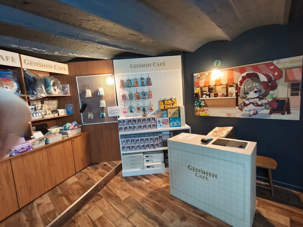 Décoration du Genshin Café avec un comptoir, une vitrine de produits dérivés et une fresque murale dans un style anime.