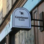 Panneau du Genshin Café suspendu à une façade parisienne, invitant les visiteurs à découvrir l'univers du jeu vidéo Genshin Impact dans un cadre convivial.