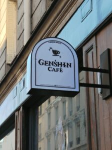 Panneau du Genshin Café suspendu à une façade parisienne, invitant les visiteurs à découvrir l'univers du jeu vidéo Genshin Impact dans un cadre convivial.