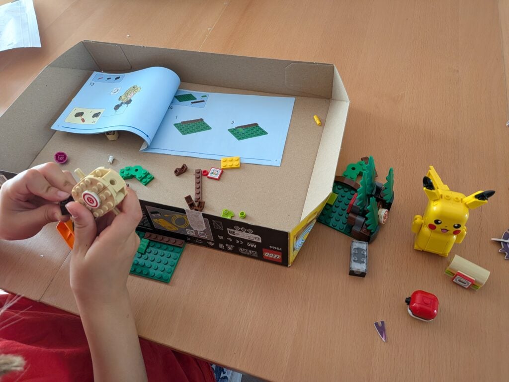Enfant construisant un LEGO Pikachu avec la boîte LEGO Pokémon 72164, entouré d'autres pièces et figurines sur une table en bois.