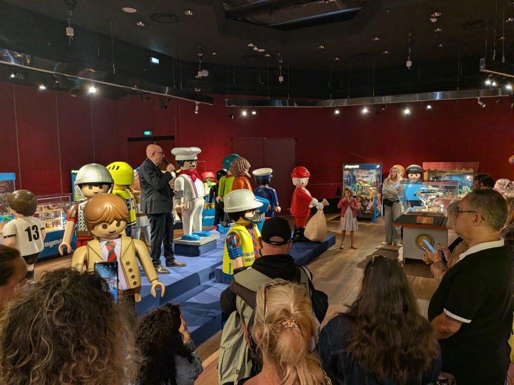 Famille et collectionneurs découvrant une exposition éphémère de figurines Playmobil à Drouot, avec des figurines en vitrine et des visiteurs admirant les modèles.