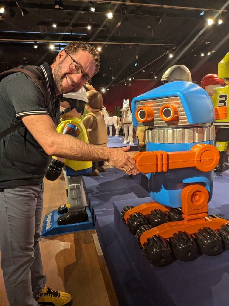 Homme souriant serrant la main d'un robot Playmobil géant lors d'une exposition, avec d'autres figurines en arrière-plan.