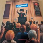 Figurines Playmobil lors d'une vente aux enchères à Drouot, avec un animateur en costume et des spectateurs numérotés.