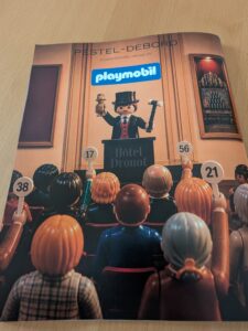 Figurines Playmobil lors d'une vente aux enchères à Drouot, avec un animateur en costume et des spectateurs numérotés.