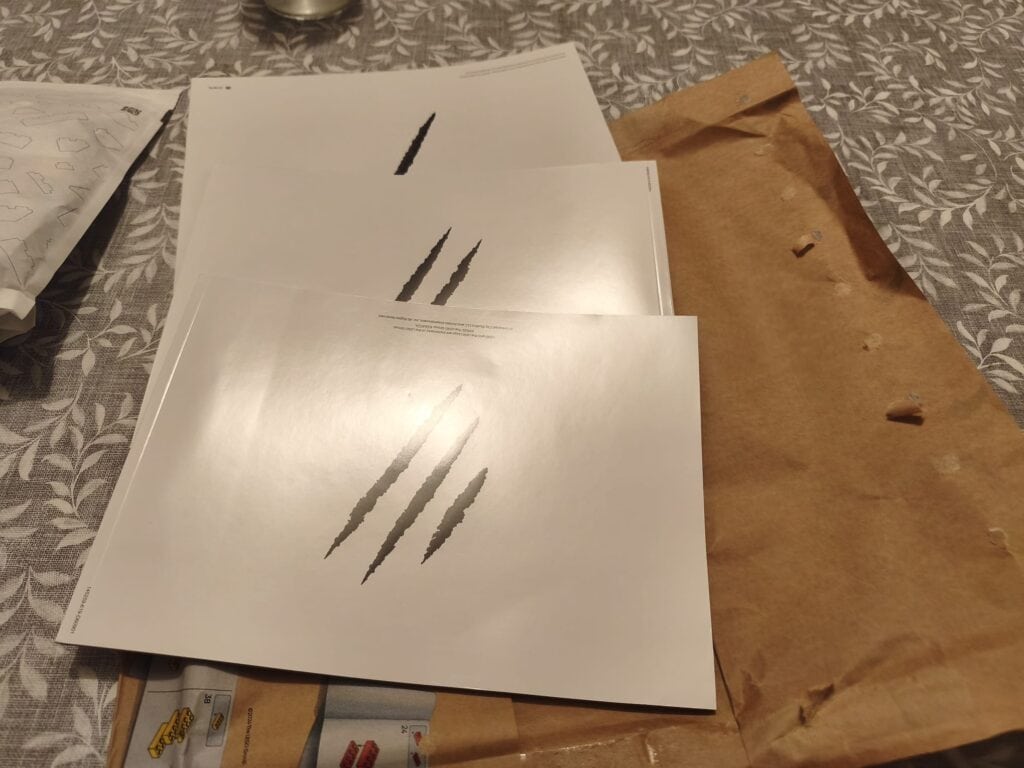 Trois feuilles de papier avec des marques de griffes de dinosaure, posées sur une table avec un sac en papier marron à côté.