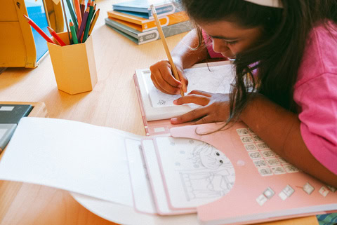 Jeune fille en train de dessiner et colorier un livre d'activités sur un bureau, entourée de fournitures scolaires colorées.
