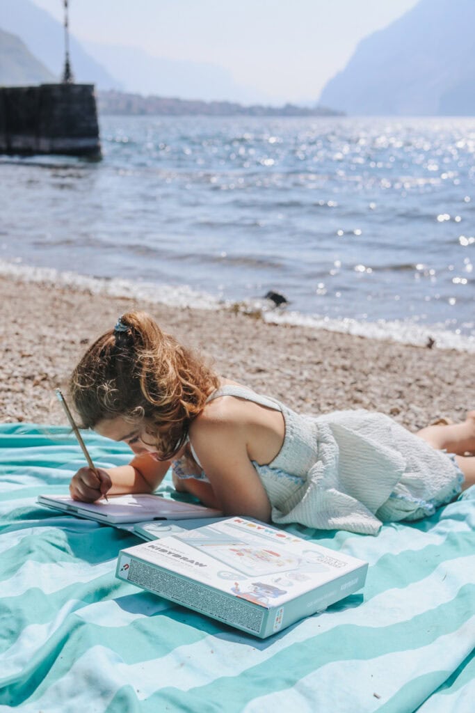 Jeune fille en robe blanche dessinant sur un carnet à la plage avec vue sur la mer et les montagnes en arrière-plan.