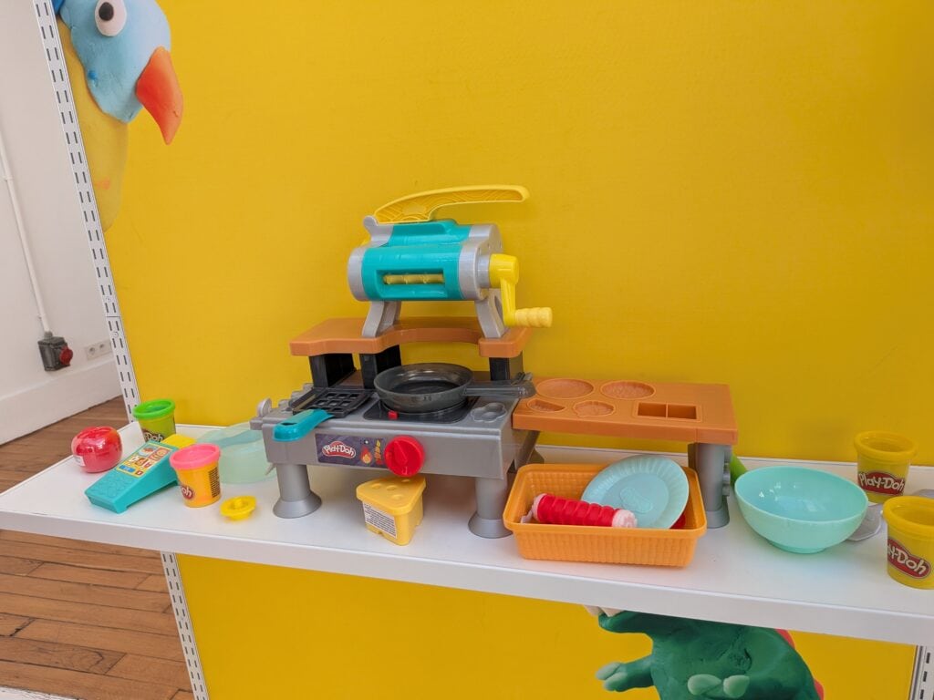 Jeu de cuisine pour enfants avec accessoires colorés et jouets en plastique sur une table blanche contre un mur jaune.