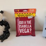Sac à dos de dossier criminel Pocket avec le titre 'Qui a tué Isabella Vega?' placé entre un robot en LEGO et une console de jeu Nintendo Game Boy.