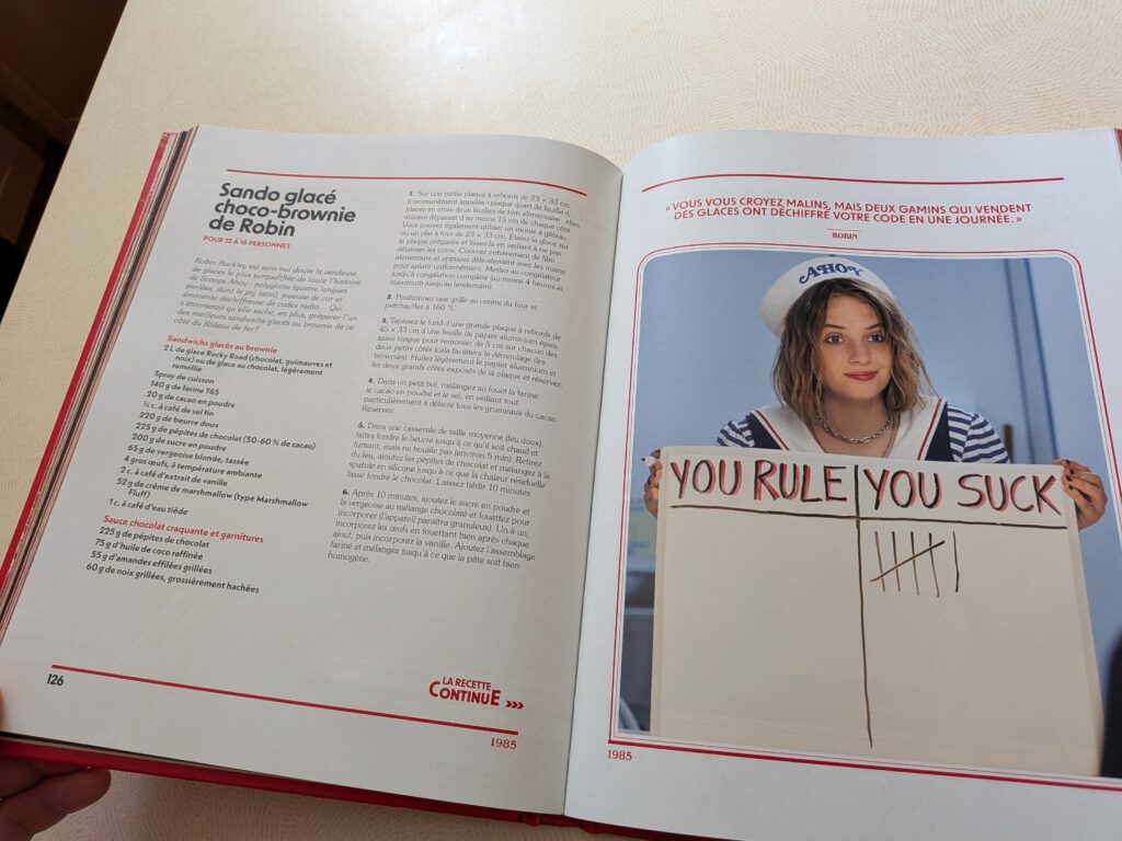 Photo d'une personne tenant un livre ouvert avec une image d'une jeune femme avec un panneau "You Rule You Suck" lors d'une activité ludique.